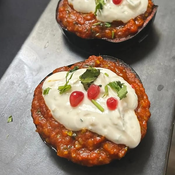 Two servings of red stew topped with white sauce, red berries, and green herbs on a dark surface.
