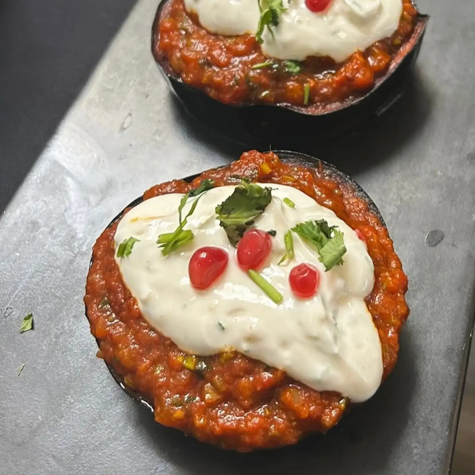 Two servings of red stew topped with white sauce, red berries, and green herbs on a dark surface.