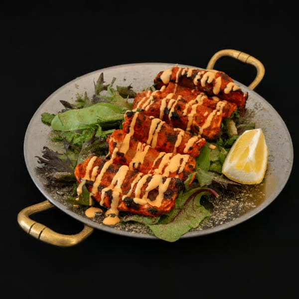 Grilled chicken tikka skewers with sauce drizzle, fresh greens, and a lemon wedge on a metal platter.